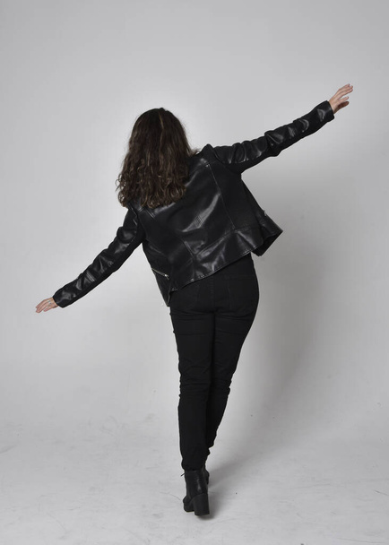 Full length portrait of young woman with natural brown hair,  wearing black leather scifi outfit with jacket, standing pose  with back to the camera, on light studio background. - Foto, Bild