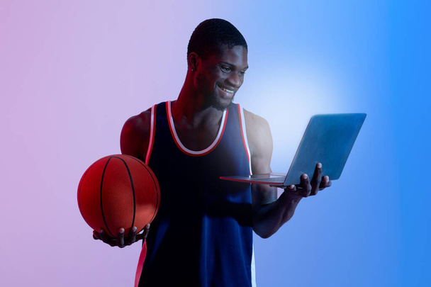 Cheerful African American sports fan holding laptop pc and basketball in neon light - Photo, image
