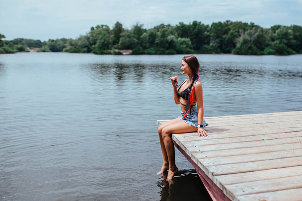 Sexy bella bruna prende il sole sul ponte vicino al lago o al fiume in costume da bagno nero. Concetto di turismo e ricreazione.  - Foto, immagini