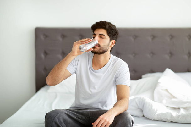 Comenzando un nuevo día con agua fresca. Hombre árabe tomando un vaso de agua sentado en la cama por la mañana - Foto, imagen