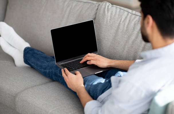 Jovem árabe cara usando laptop moderno com tela preta vazia em casa, eclining no sofá, mockup, sobre tiro no ombro - Foto, Imagem