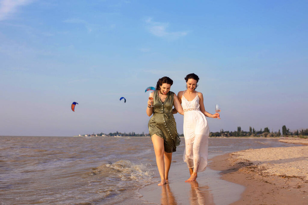 Felices amigas adultas en traje de verano sonriendo mientras disfrutan del vino en la playa de arena cerca del mar - Foto, imagen