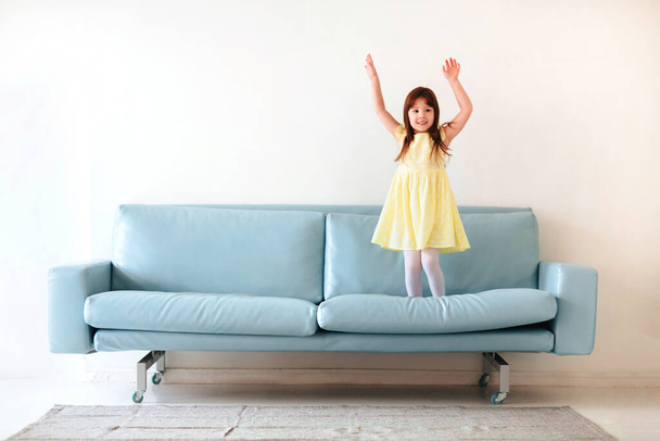 Little caucasian cute lovely girl in yellow dress playing and jumping on blue sofa in living room at home isolated on white concrete wall background. Childhood and leisure time concept - Photo, Image