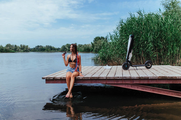 Eine schöne Brünette im Badeanzug sonnt sich auf einer Holzbrücke und in der Nähe steht ein Motorroller. Ökologie und Gesundheitswesen. Hochwertiges Foto - Foto, Bild