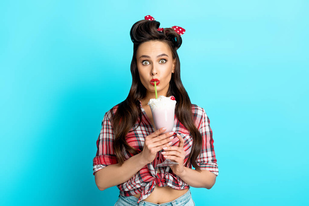 Schockierte junge Pinup-Frau in Retro-Klamotten trinkt leckeren Milchshake auf blauem Studiohintergrund - Foto, Bild