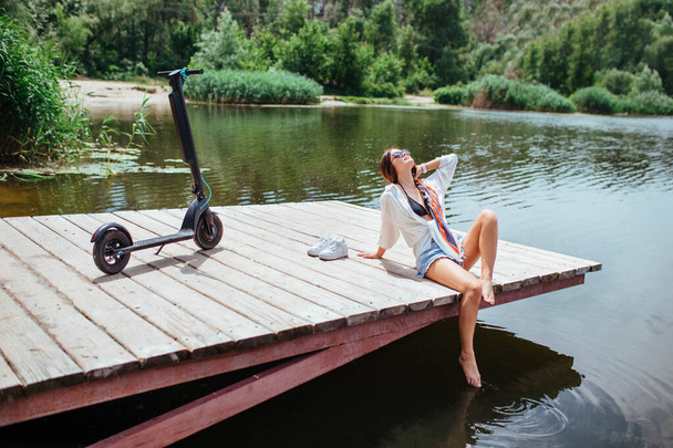 A beautiful brunette girl rests on a wooden bridge near the river next to an electric scooter. The concept of ecology and electric transport. - Photo, Image