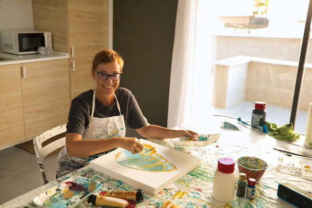 Mujer madura con gafas pintando un cuadro de una mariposa en su estudio de pintura mirando a la cámara. - Foto, Imagen