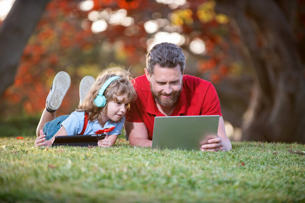 ευτυχισμένη οικογένεια του μπαμπά και του γιου χρήση φορητού υπολογιστή και ακουστικά στο πάρκο, οικογενειακό Σαββατοκύριακο. - Φωτογραφία, εικόνα