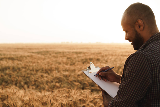 Man making notes on clipboard while standing in wheat field - Fotografie, Obrázek