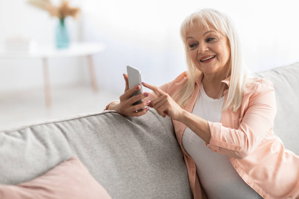 Usmívající se starší žena pomocí mobilního telefonu odpočívá na pohovce - Fotografie, Obrázek
