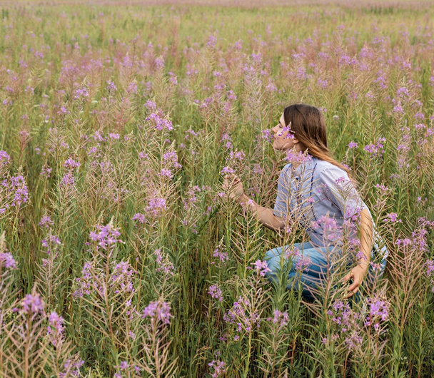 Beautiful smiling blond young woman in light purple shirt sitting on field of fireweed flowers sniffing the scent of pink flower copy space enjoing the nature - Photo, Image