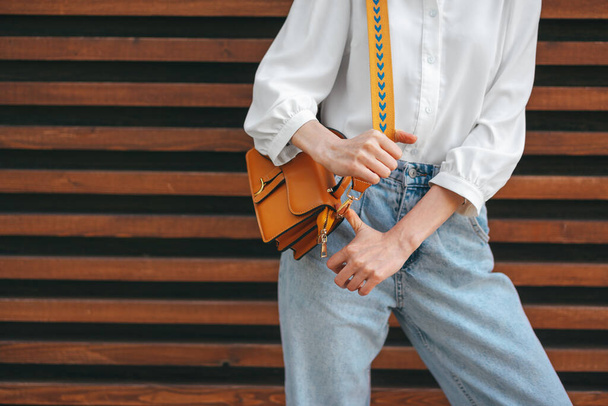 Fashion photo of woman wearing white shirt, jeans and brown crossbody bag against wooden background - Zdjęcie, obraz