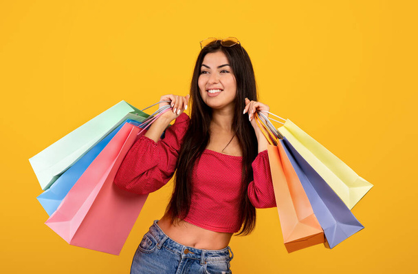 Seasonal sales. Portrait of happy armenia woman holding colorful shopping bags with purchases, yellow background - Foto, imagen