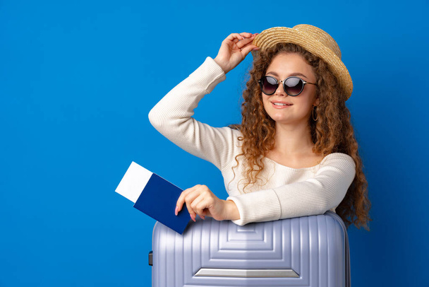 Young beautiful woman in hat on vacation with suitcase against blue background - Фото, зображення