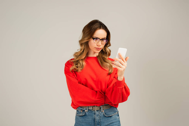 mujer disgustada con gafas de vista usando smartphone aislado en gris - Foto, imagen