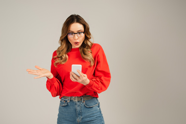 mujer sorprendida en gafas de vista mirando teléfono inteligente aislado en gris - Foto, imagen