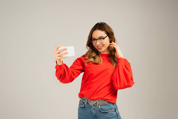 positive woman in eyeglasses taking selfie on smartphone isolated on grey - Foto, afbeelding