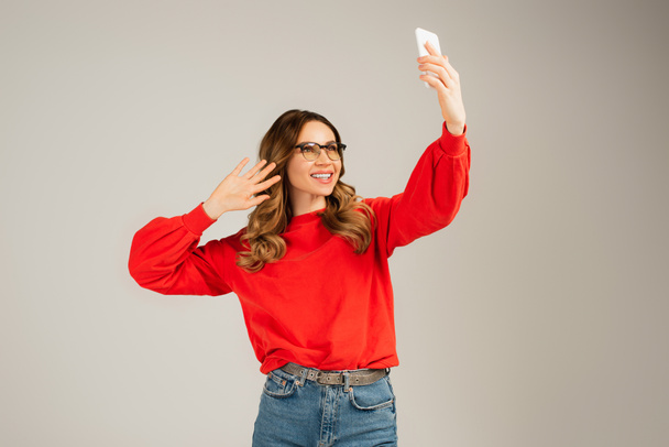 glückliche Frau in Brille und Sweatshirt mit Videoanruf auf Smartphone isoliert auf grau - Foto, Bild