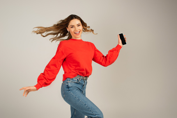 excited woman in wireless earphones holding smartphone with blank screen isolated on grey  - Foto, Imagem