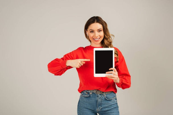 cheerful woman in wireless earphones pointing at digital tablet with blank screen isolated on grey  - Φωτογραφία, εικόνα