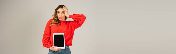 Gri pankartta izole edilmiş boş ekranlı dijital tableti tutan şok olmuş bir kadın. - Fotoğraf, Görsel