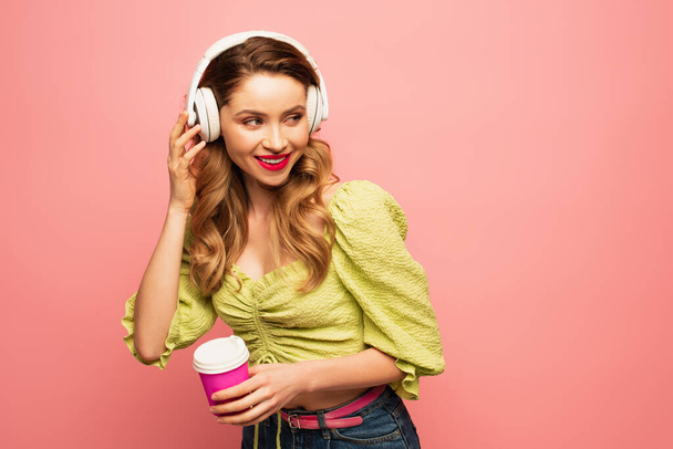 χαρούμενη γυναίκα ρυθμίζοντας ασύρματα ακουστικά και κρατώντας χάρτινο κύπελλο απομονωμένο σε ροζ - Φωτογραφία, εικόνα