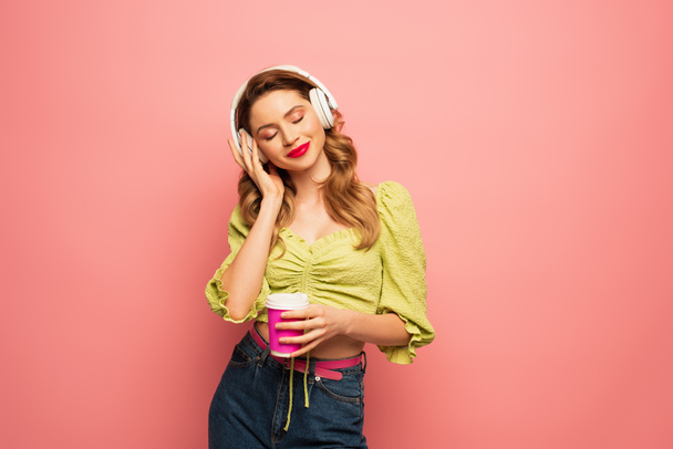 ευχαριστημένη γυναίκα ρυθμίζοντας ασύρματα ακουστικά και κρατώντας χάρτινο κύπελλο απομονωμένο σε ροζ - Φωτογραφία, εικόνα