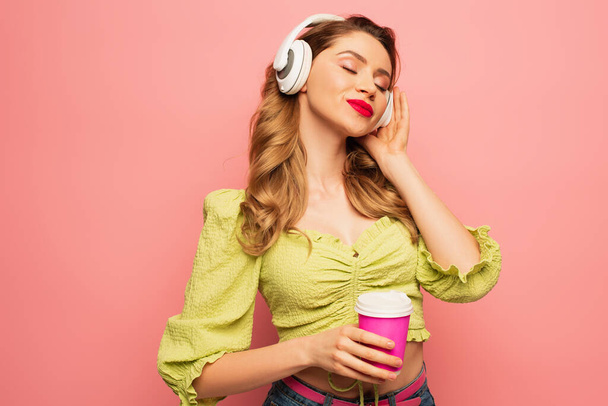 mulher feliz ajustando fones de ouvido sem fio e segurando café para ir isolado em rosa - Foto, Imagem