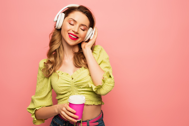 pleased woman adjusting wireless headphones and holding coffee to go isolated on pink - Photo, image