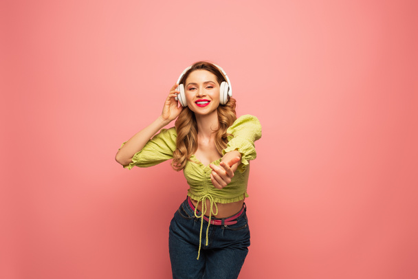 happy woman adjusting wireless headphones and standing with outstretched hand on pink - Φωτογραφία, εικόνα
