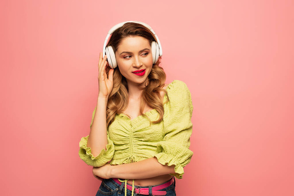 sly woman adjusting wireless headphones and looking away isolated on pink - Photo, Image