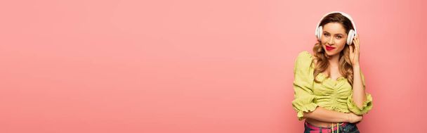 femme rusée ajustant écouteurs sans fil et regardant la caméra isolée sur rose, bannière - Photo, image