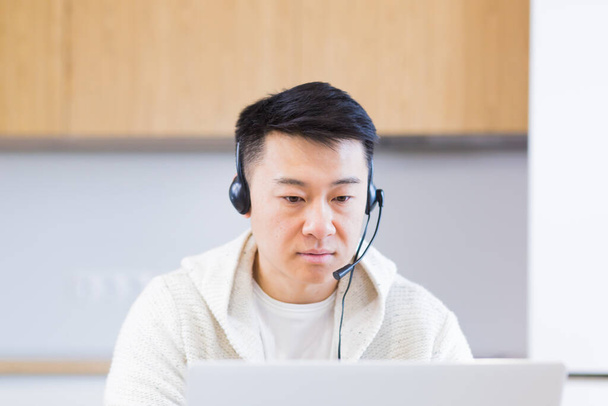 young asian man freelancer sitting at home at office desk with headset and talking online on video link via laptop in room or kitchen. The male indoors in casual clothes does the work and talks - Zdjęcie, obraz