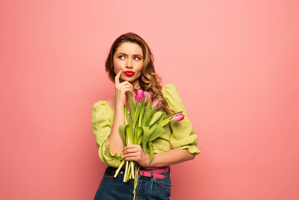 pensive woman pouting lips and holding bouquet of tulips isolated on pink - Fotoğraf, Görsel