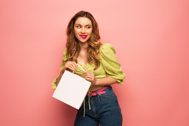 curious woman in green blouse holding shopping bag and biting lips isolated on pink  - Photo, Image