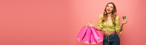 amazed woman holding credit card and shopping bags isolated on pink, banner - Fotografie, Obrázek