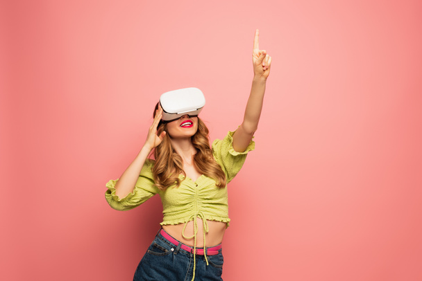 happy woman in vr headset pointing with finger isolated on pink  - Fotó, kép