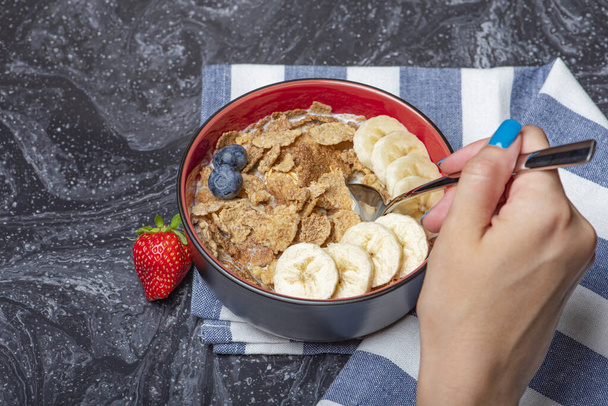 Мюсли. Завтрак, здоровое питание и диета. Мюсли с молоком и фруктами в тарелке на черном мраморном верху. Женская рука с ложкой - Фото, изображение