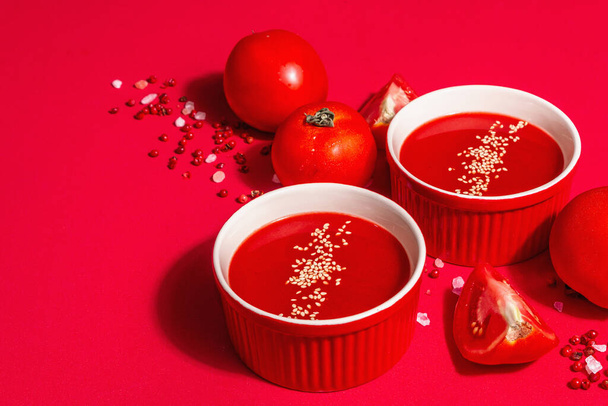 Tomatensuppe mit Sesam in Schalen auf rotem Hintergrund. Reifes Gemüse, rosa Himalaya-Salz, rosa Pfefferkorn. Ein modernes hartes Licht, dunkler Schatten, Kopierraum - Foto, Bild