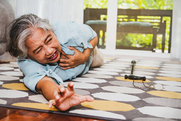 Starsza Azjatka samotnie w domu, cierpiąca na atak serca, upada na ziemię, okulary i laska zsuwają się w bólu, szukając pomocy: Koncepcja opieki nad starszym ubezpieczeniem zdrowotnym - Zdjęcie, obraz