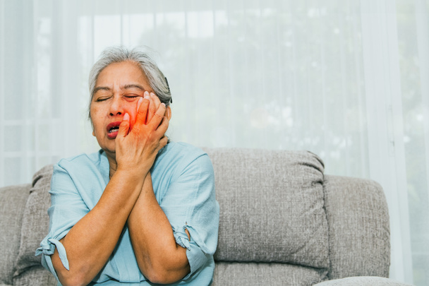 Ältere Frau, die zu Hause auf dem Sofa sitzt, hat Zahnschmerzen, Hohlräume, plötzliche Zahnfleischentzündung oder Zahnempfindlichkeit durch Degeneration, Alterung und Mundschmutz: Krankenversicherungskonzept - Foto, Bild