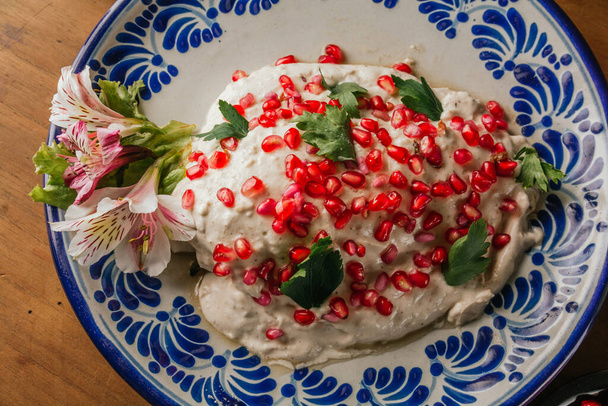 Chiles en Nogada, 100% мексиканский рецепт с его основными ингредиентами с одной стороны, такими как грецкий орех, красная градана, служил на тарелке талавера Пуэбла, на деревянном столе. - Фото, изображение