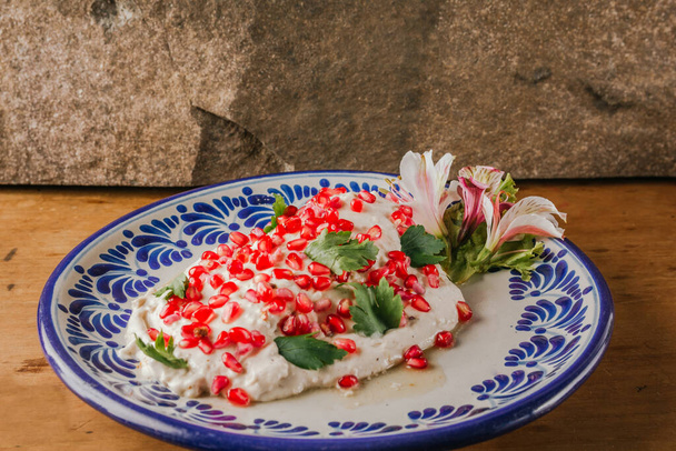 Chiles en Nogada, ein 100% mexikanisches Rezept mit seinen Hauptzutaten auf der einen Seite, wie Walnuss, rote Gradana, serviert auf einem Teller aus Puebla Talavera, auf einem Holztisch. - Foto, Bild