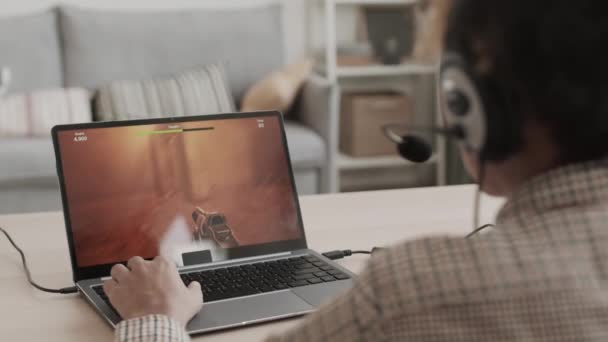 Pohled přes rameno na nepoznatelnou osobu sedící za stolem ve svém pokoji, hrající střílečku na přenosném počítači - Záběry, video