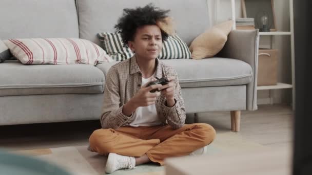 Πλήρης κλείδωμα του δεκατριάχρονου αγοριού που κάθεται στο πάτωμα δίπλα στον καναπέ στο σπίτι, κρατώντας χειριστήριο κονσόλας παιχνιδιών, παίζοντας και φεύγοντας αφού απέτυχε - Πλάνα, βίντεο