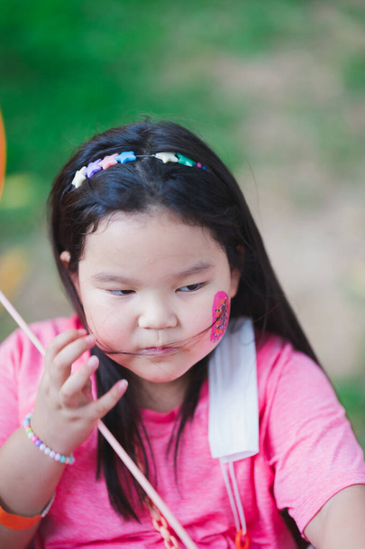 Vertical image. Asian girl sits to rest from Easter activities. Children paint watercolor cartoons on cheeks. Child decorate their hair with star headband. Kid 6-7 years old wear pink t-shirts. - Photo, Image