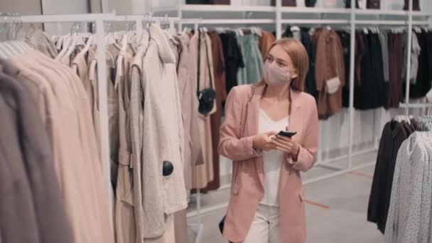 Yüz maskeli genç bir kadının cep telefonuyla yazı yazarken ve kıyafet mağazasında alışveriş yaparken çekilmiş yavaş bir resmi. Elbise askılığından kazağını çıkarıp uzaklaşıyor. - Video, Çekim