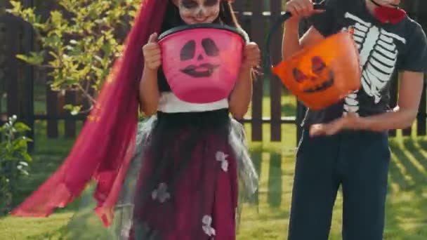 Portrait incliné de la petite fille et du garçon en costumes d'Halloween tenant des paniers pour tricher ou traiter et posant pour la caméra à l'extérieur le jour ensoleillé - Séquence, vidéo
