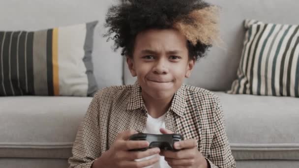 Στήθος δεκατρία ετών Αφρο αγόρι κάθεται στο πάτωμα δίπλα στον καναπέ στο σπίτι, κρατώντας χειριστήριο κονσόλας παιχνιδιών, παίζει και ενθουσιάζεται με τη νίκη - Πλάνα, βίντεο
