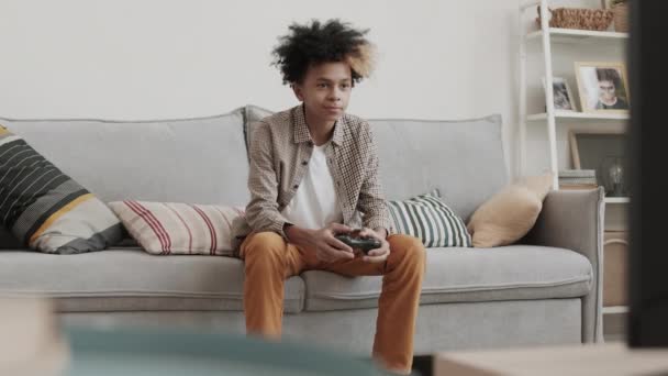 Μέτριο μήκος δεκατριάχρονου αγοριού που κάθεται στον καναπέ στο σπίτι, κρατώντας χειριστήριο κονσόλας παιχνιδιών, παίζοντας - Πλάνα, βίντεο